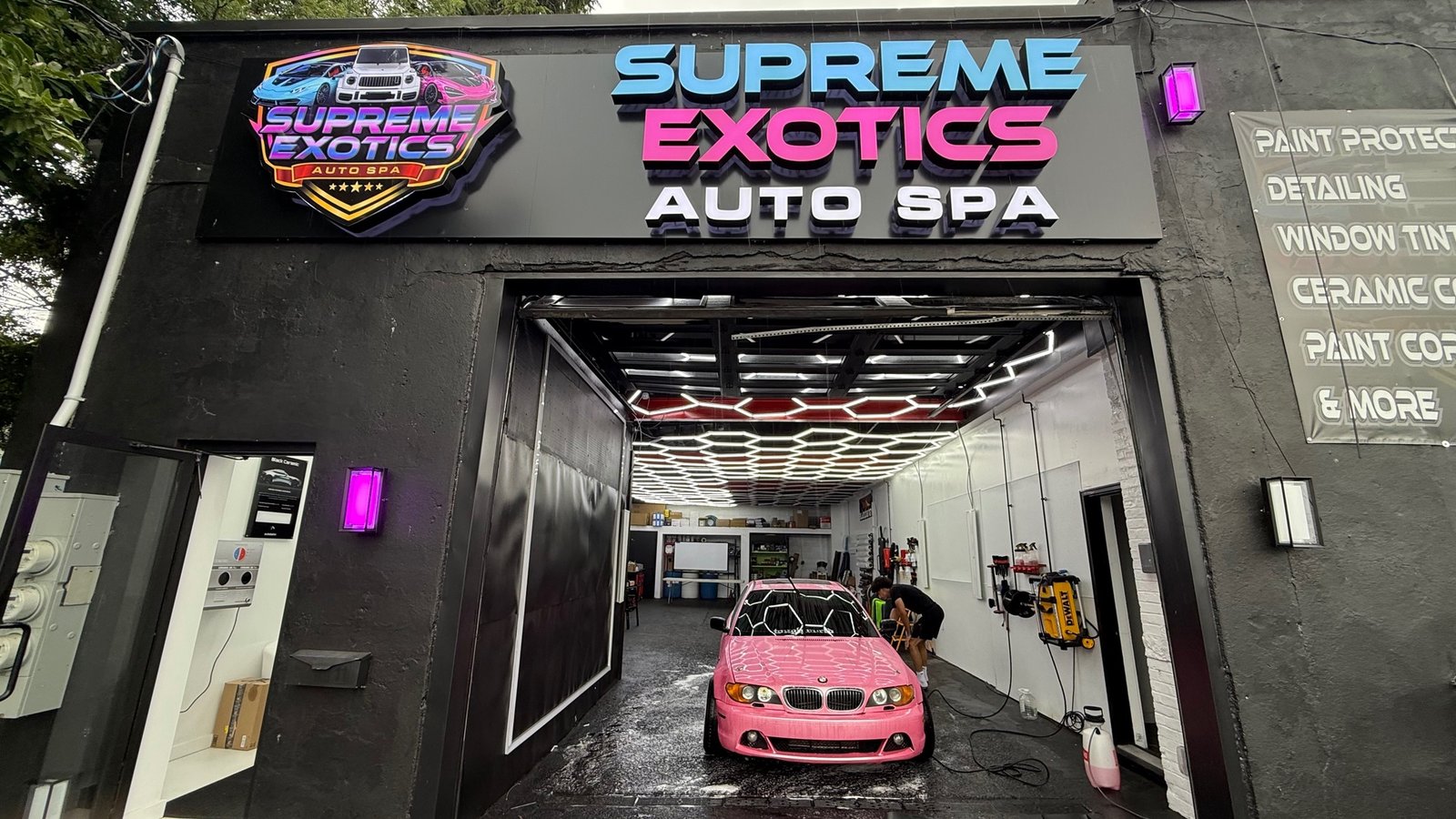 The storefront in daylight — shield sign and channel letters above the open bay, hex ceiling lights running back inside, a car mid-wash