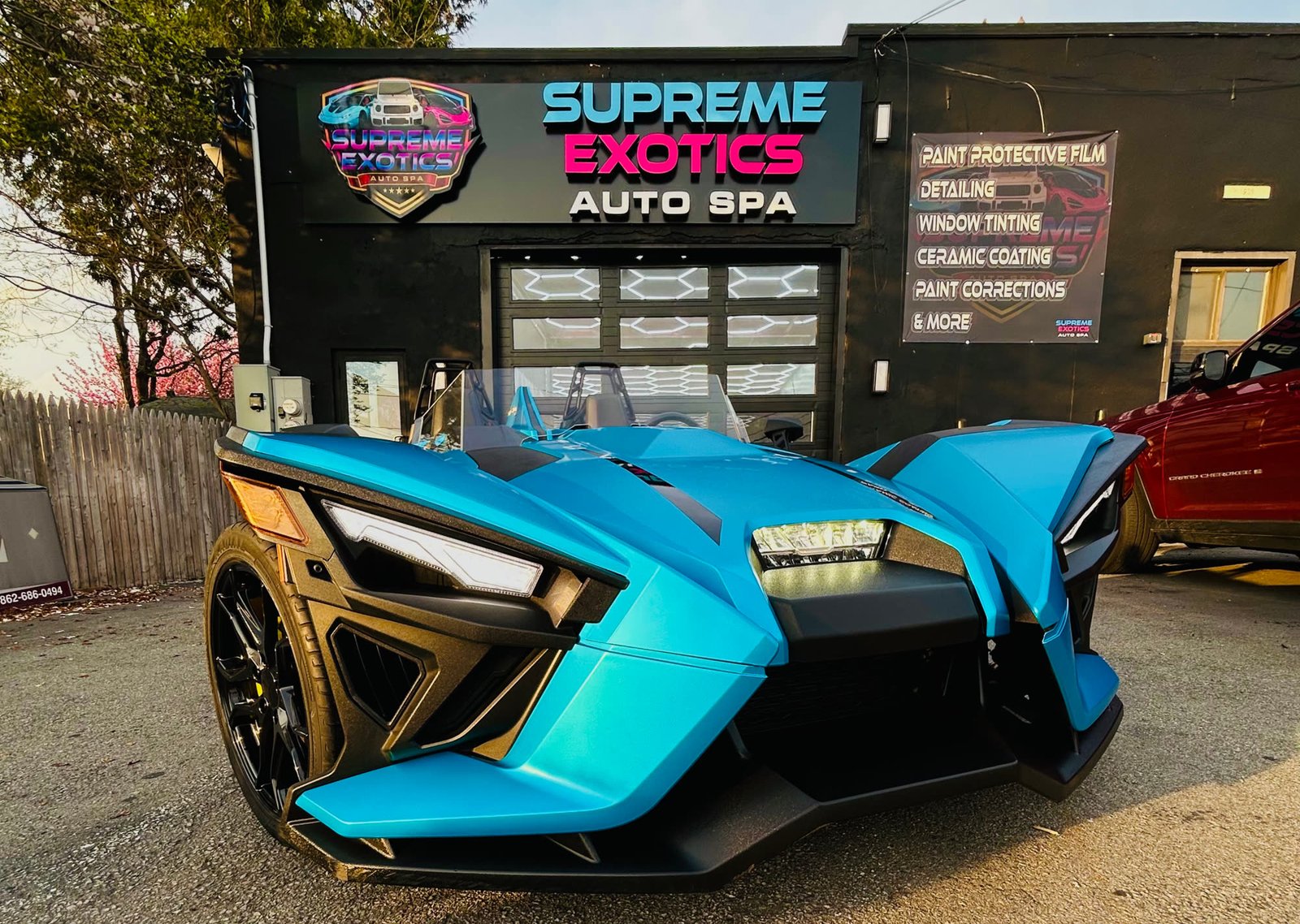 A bright blue Polaris Slingshot parked outside the shop at golden hour, the Supreme Exotics sign and service list on the wall behind it